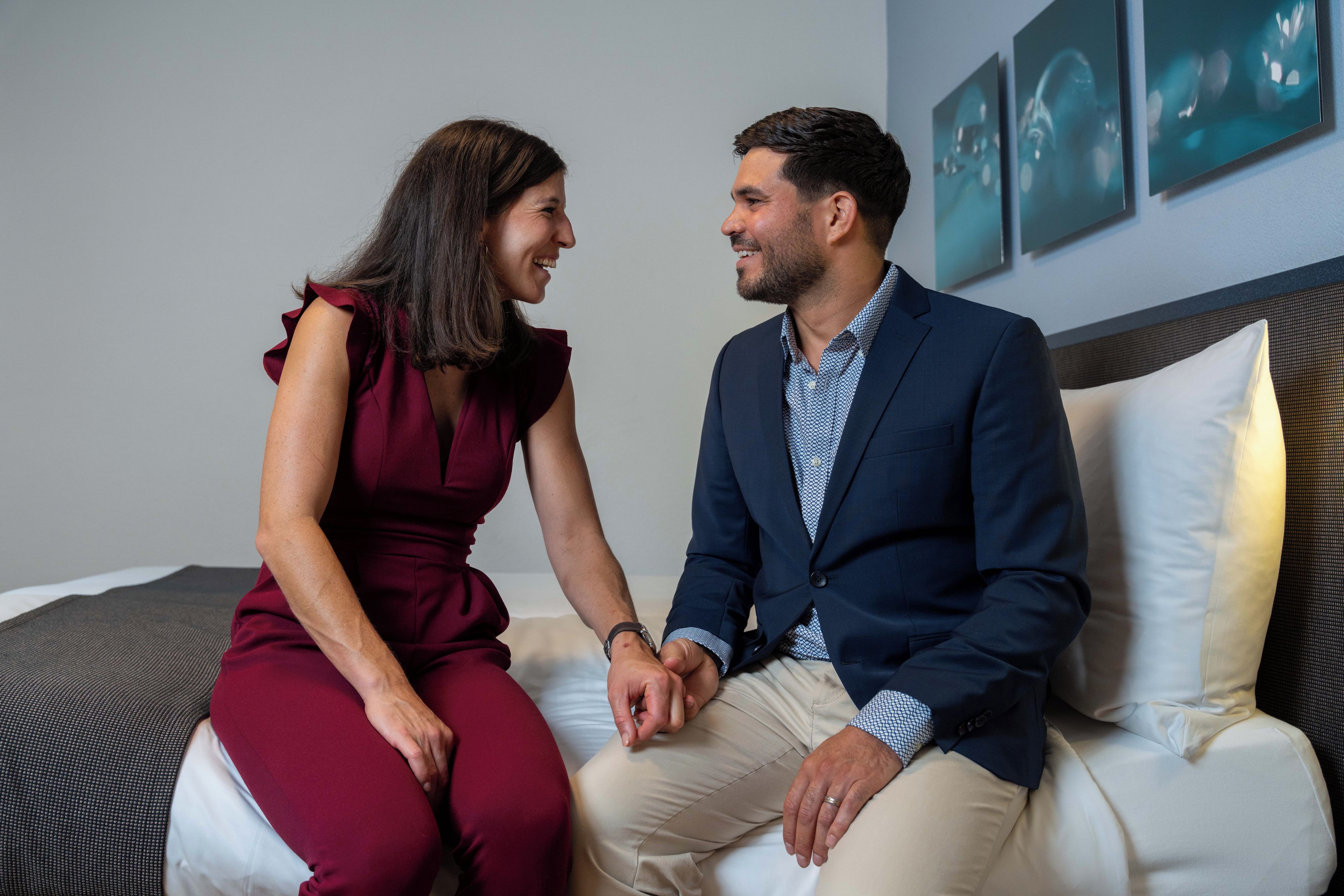 A couple seated on a bed with a headboard, in a room with a plain wall and a framed picture.