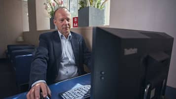 Ein Mann sitzt an einem Schreibtisch mit Computer, Tastatur und Maus. Im Hintergrund ist ein Fenster mit Blumen zu sehen.