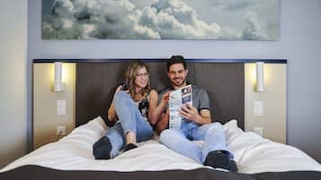 A couple sitting on a bed with a headboard, holding a magazine, in a room with a wall-mounted light fixture and a painting of clouds.