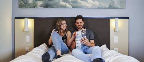 Una pareja sentada en una cama con cabecera, sosteniendo una revista, en una habitación con una lámpara de pared y un cuadro de nubes.