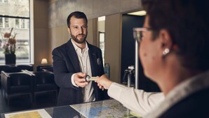 A man in a suit handing over a card to another person at a reception desk with a map and a schedule on the counter. - Bristol Hotel (Zürich)