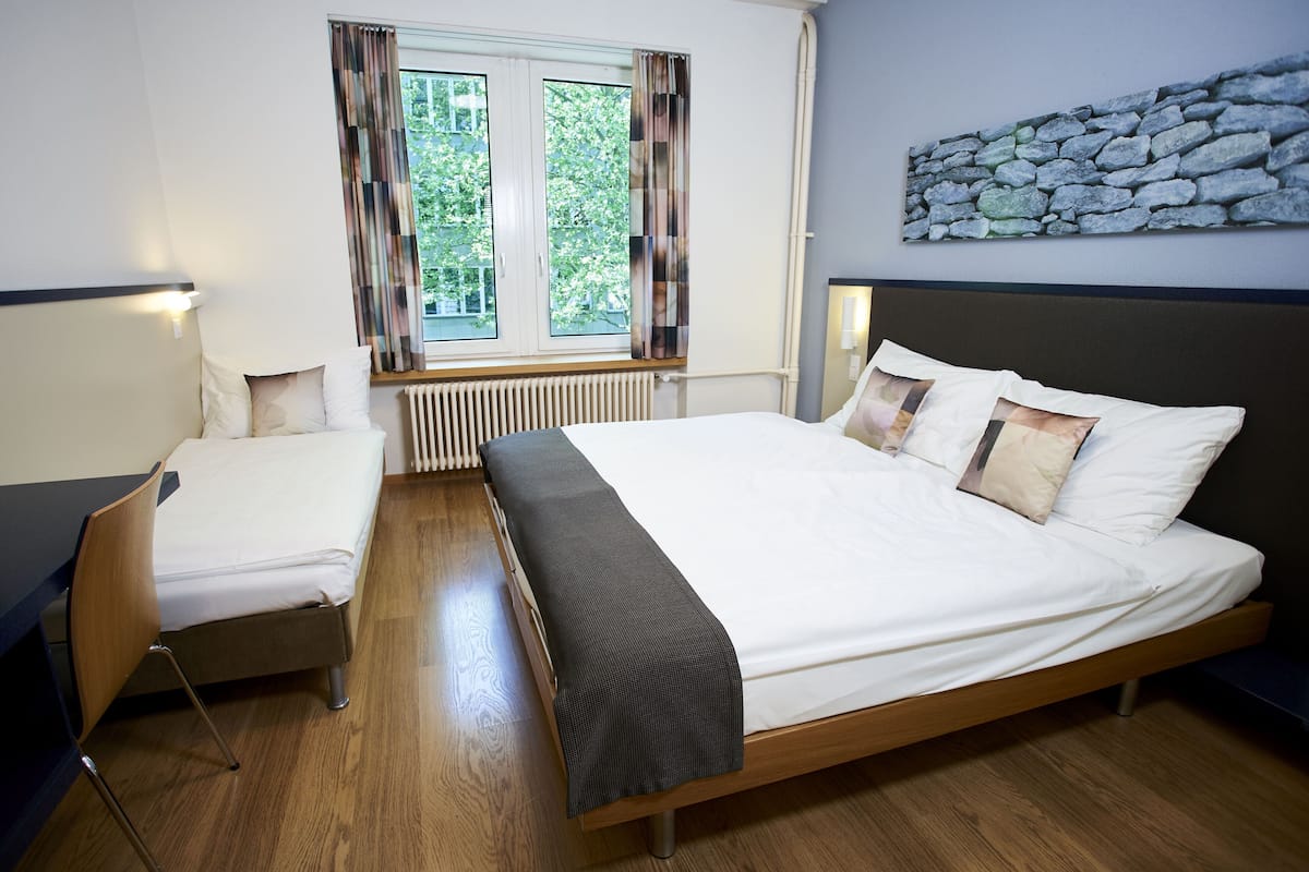 Une chambre avec un lit, une chaise, une fenêtre avec des rideaux, un radiateur et une œuvre d’art murale en pierre.