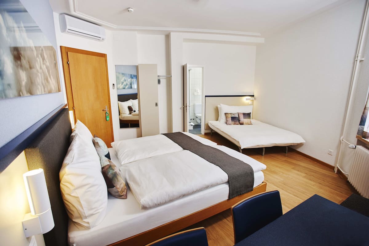 Une chambre d’hôtel avec un grand lit, un plus petit lit, un bureau et une chaise. On aperçoit une salle de bain au fond de la pièce.