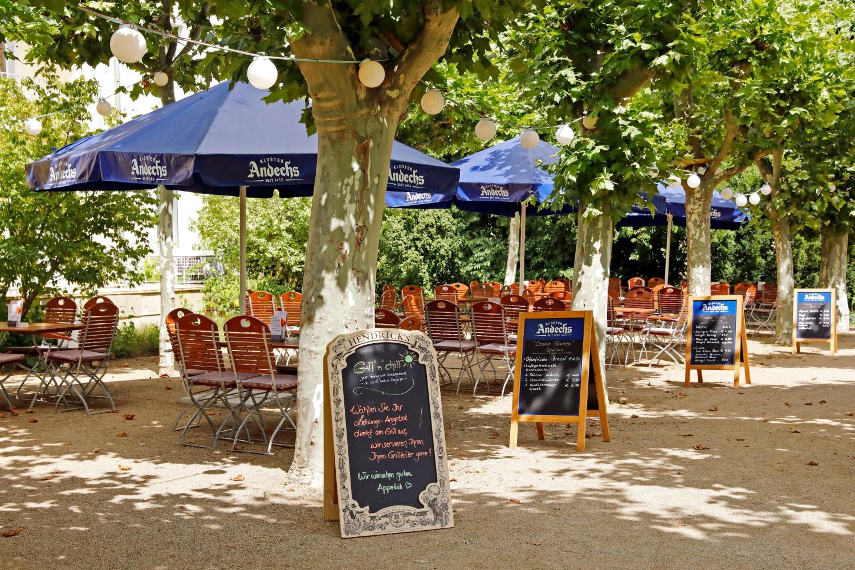 Een terras met tafels en stoelen onder blauwe parasols, met menukaarten op standaardjes.