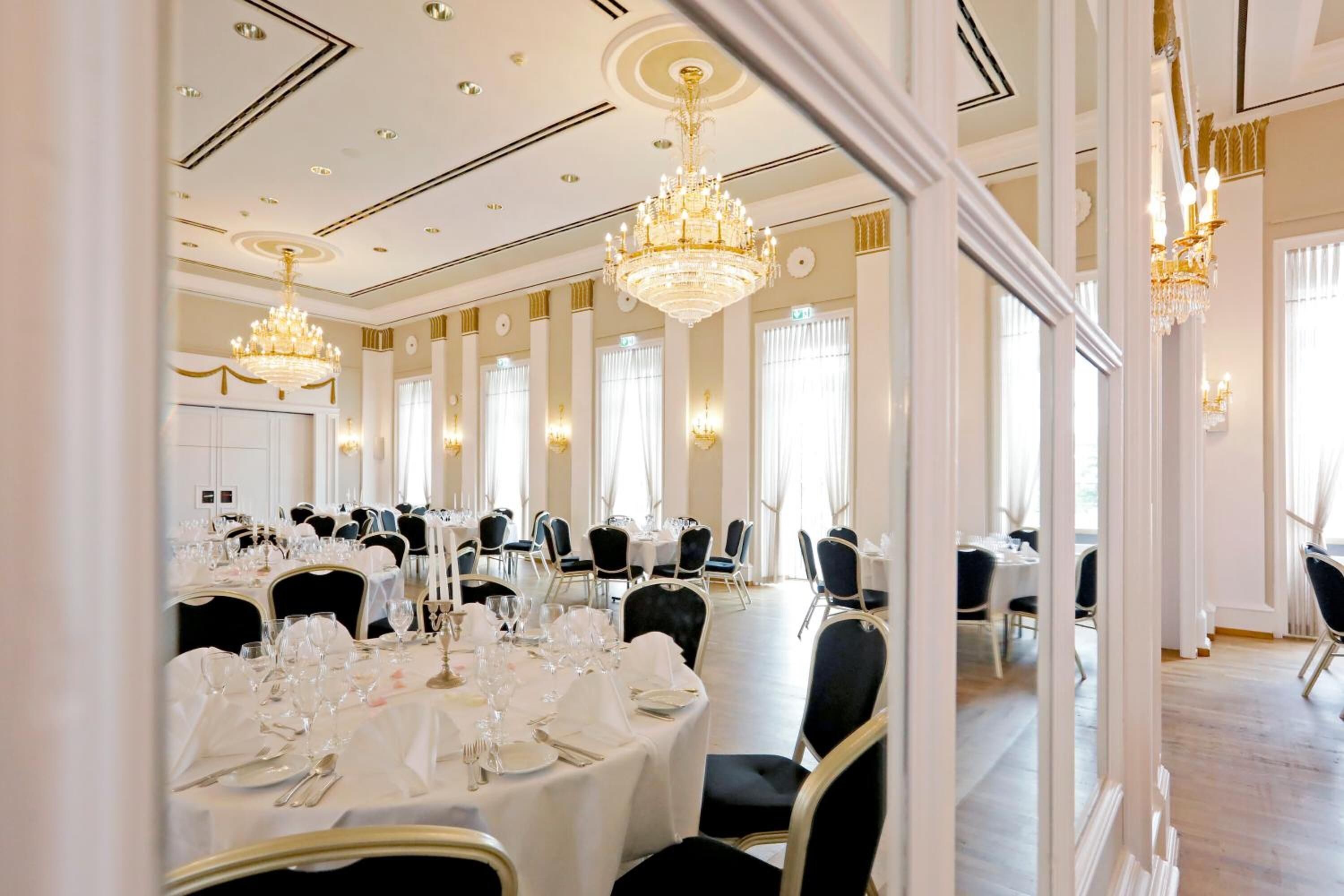 A formal dining room with multiple round tables set for a meal, large chandeliers, and tall windows.