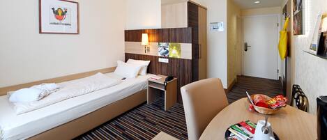A hotel room with a single bed, a desk with a lamp, a dining table with breakfast items, and a framed picture on the wall.