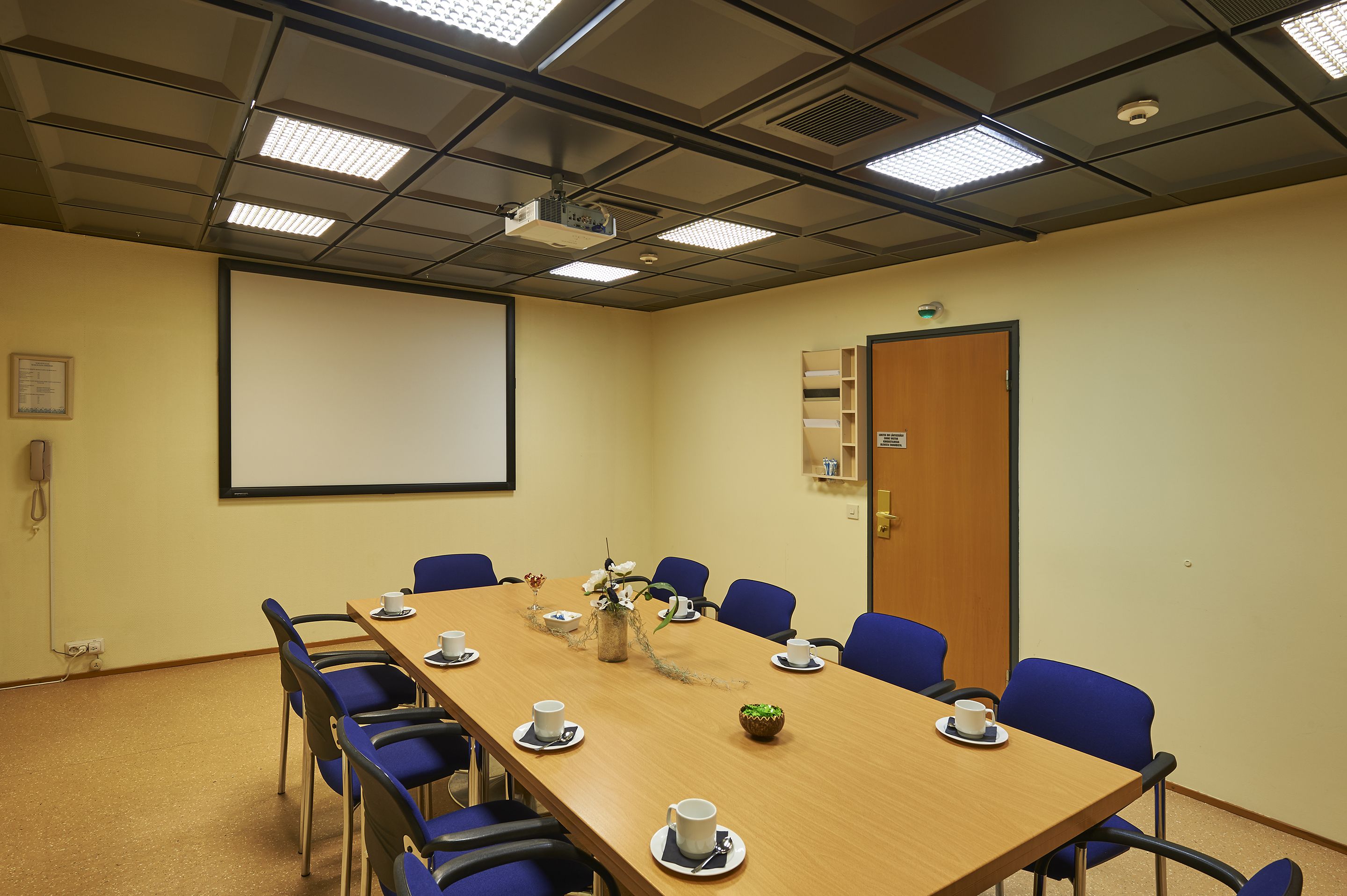 A conference room with a long table, blue chairs, a projector screen, and a door.