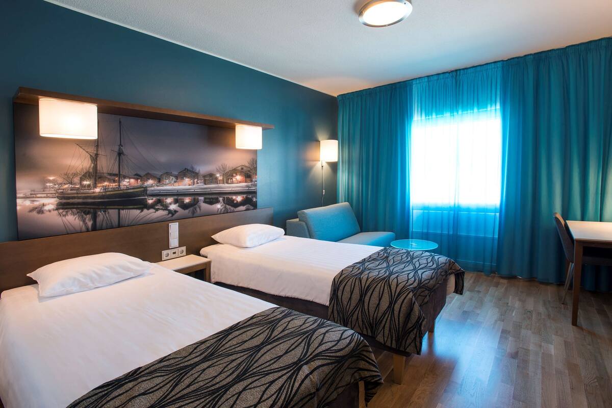 Habitación de hotel con dos camas, un escritorio, una silla, un ventanal con cortinas y una lámpara de pared.
