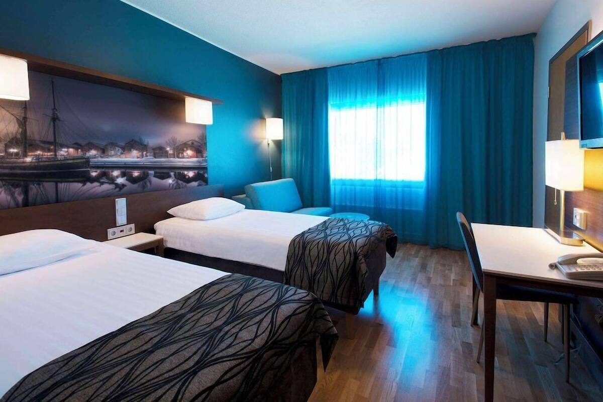 Habitación de hotel con dos camas, un escritorio, una silla, un televisor y un ventanal con cortinas.