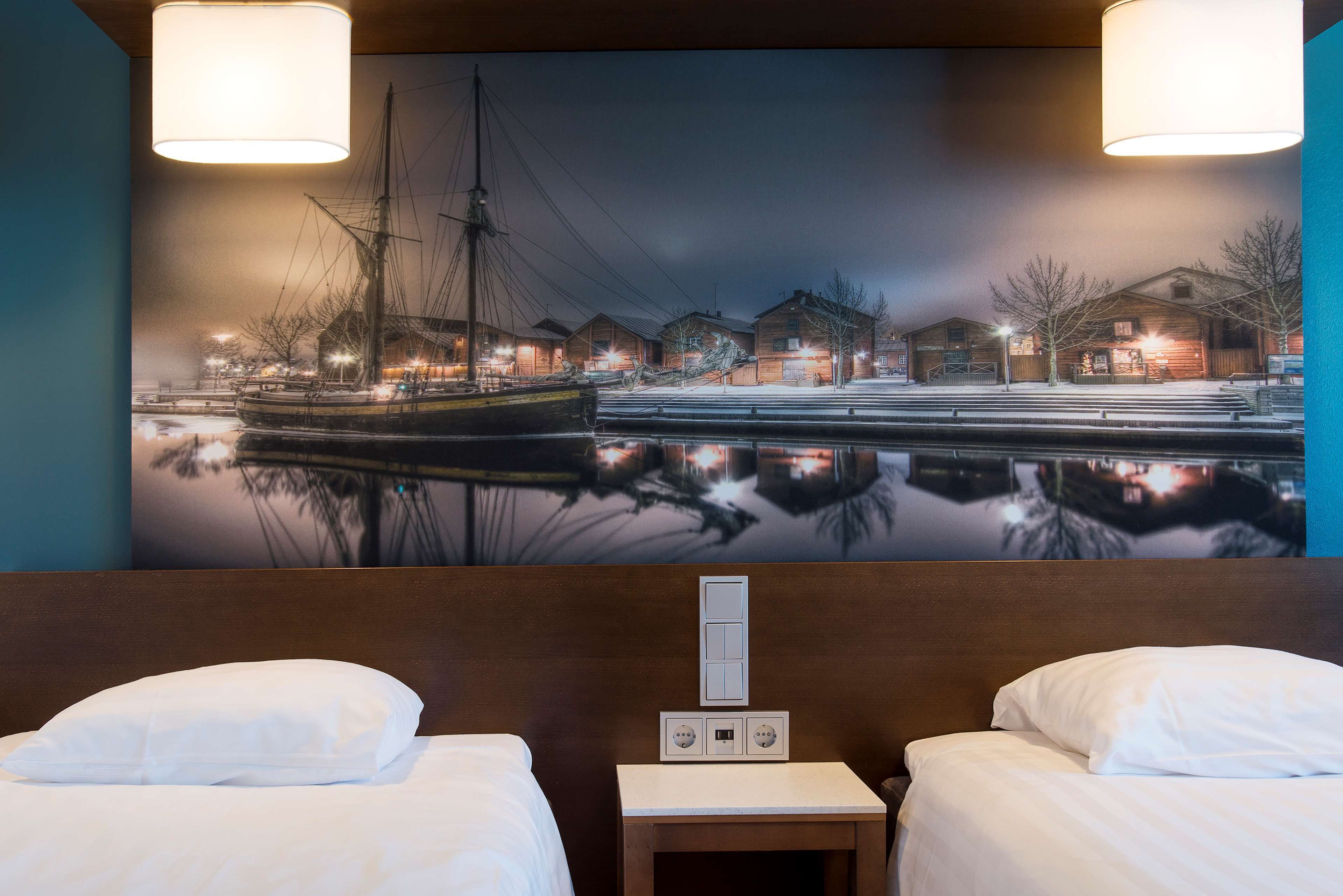 Habitació d'hotel amb dos llits, una tauleta de nit i un mural a la paret que representa una escena costanera.