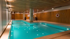 Une piscine intérieure aux murs et au plafond recouverts de carreaux, une bouée de sauvetage fixée au mur et un espace d’accueil avec un mur en panneaux de bois.