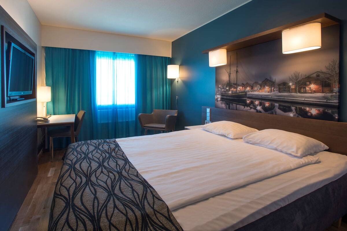 Habitación de hotel con una cama grande, un escritorio con silla, un televisor y un cuadro en la pared.