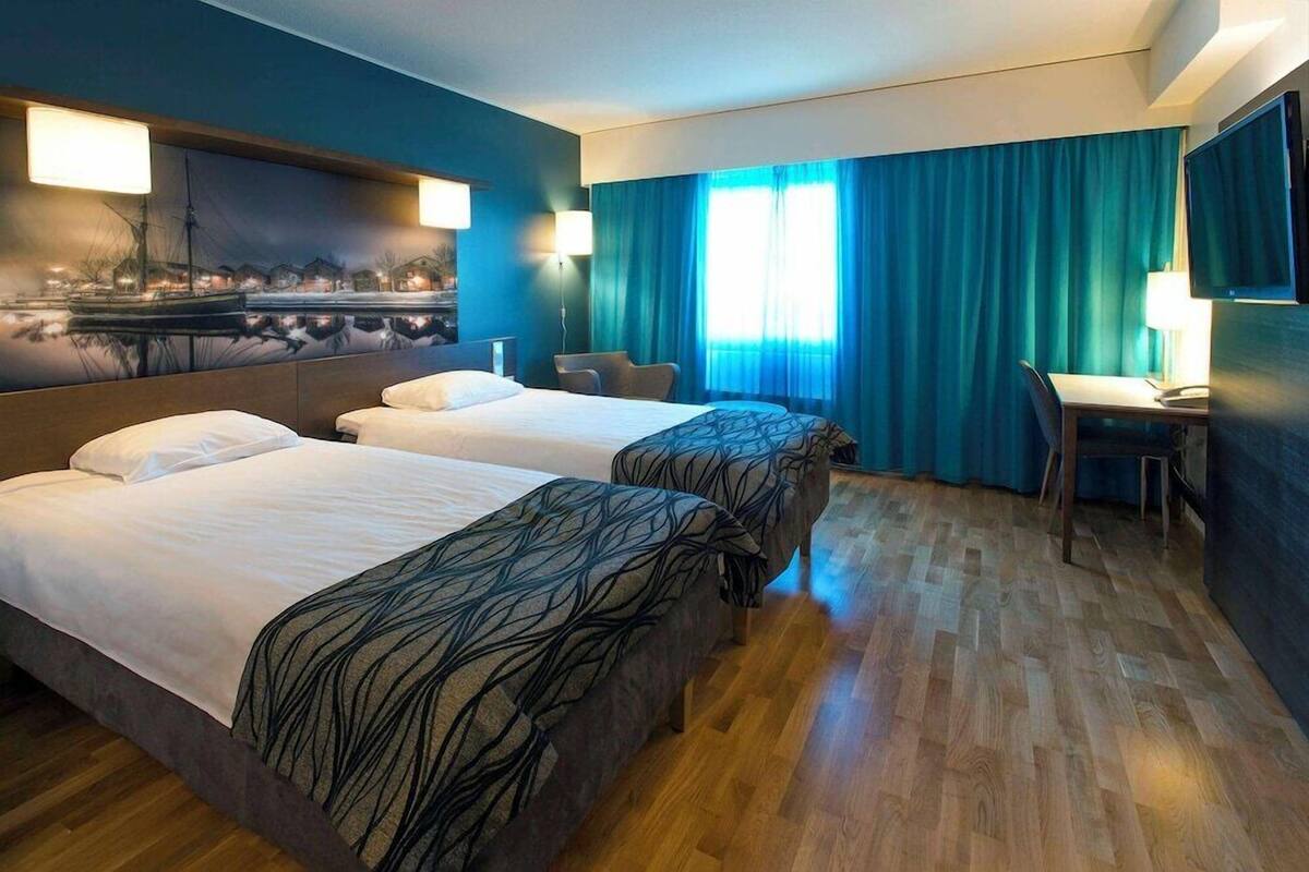 Habitación de hotel con dos camas, un escritorio, una silla y un amplio ventanal con cortinas.