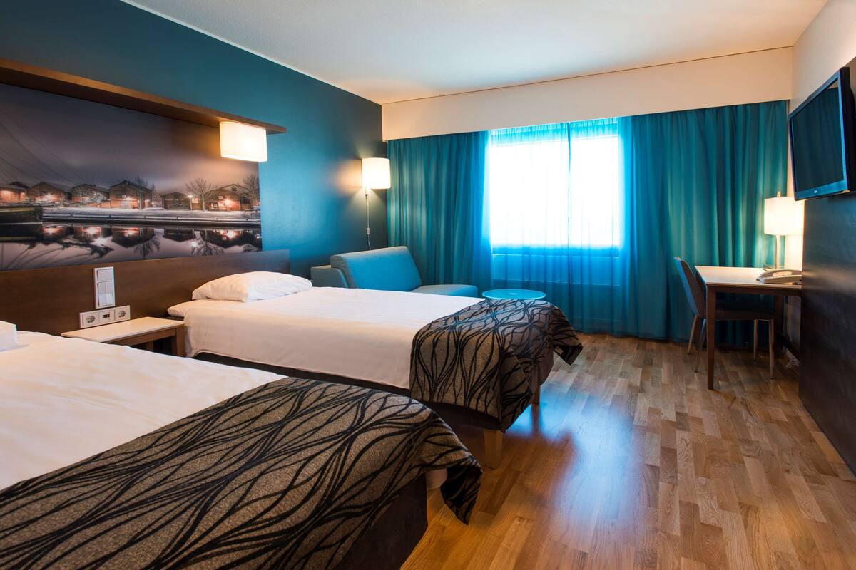 Habitación de hotel con dos camas, un escritorio, una silla, un televisor y una ventana con cortinas.