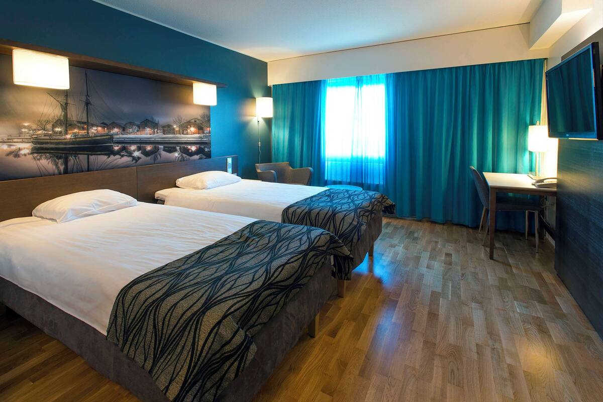 Habitación de hotel con dos camas, un escritorio, una silla, un televisor y un ventanal grande con cortinas.