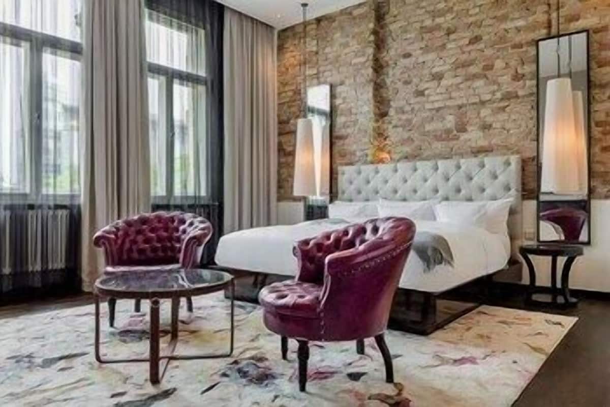 A hotel room with a large bed, two armchairs, a brick wall, and a large window.