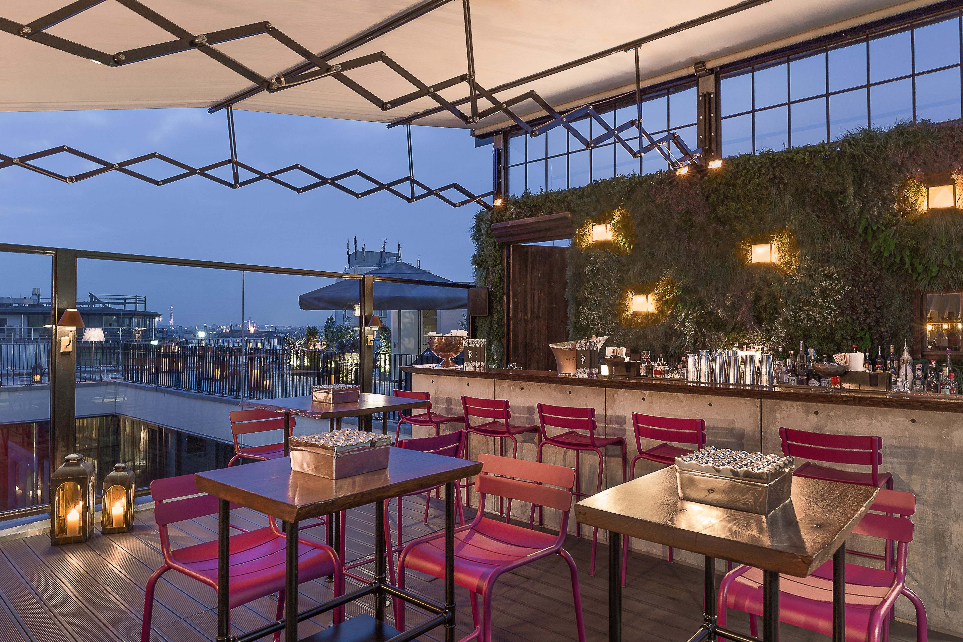 Un bar en la azotea con mesas y sillas, una barra con botellas y vistas a la ciudad.