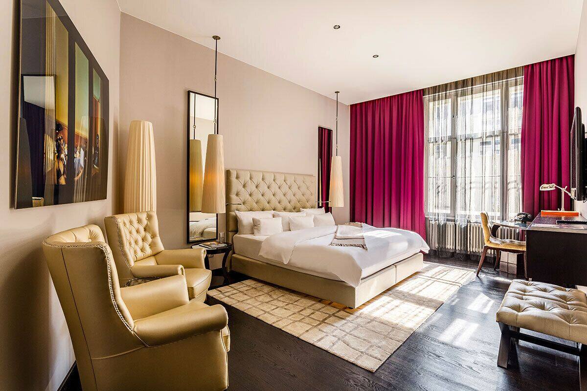 A modern hotel room with a large bed, a sitting area with a chair, a desk, and a television.
