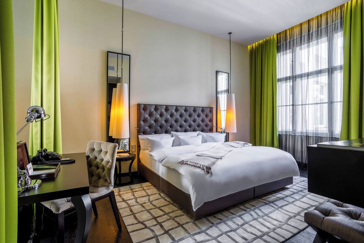 A hotel room with a large bed, a desk with a chair, a mirror, and a window with curtains.