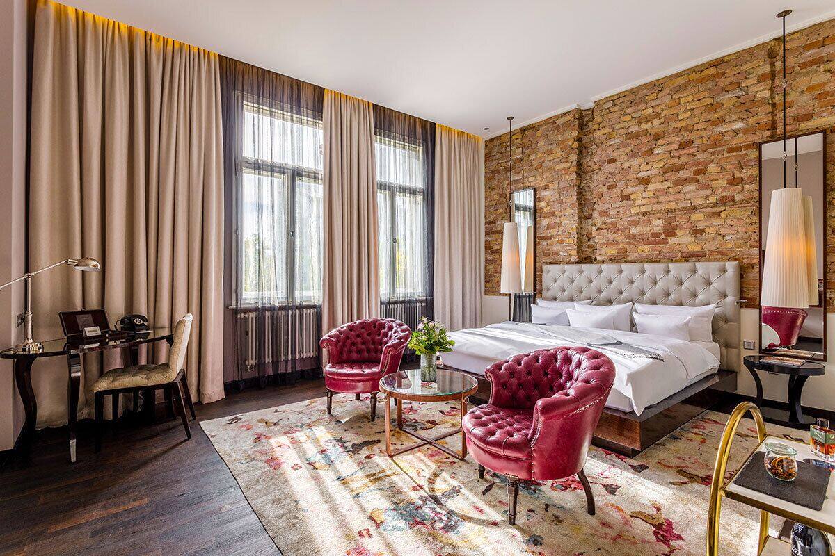 A hotel room with a large bed, two red armchairs, a small table, a desk with a lamp, and a brick wall.