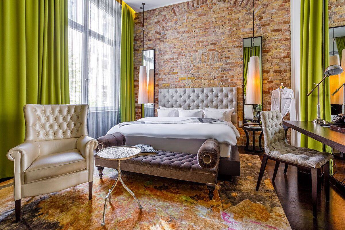 A hotel room with a brick wall, a large bed, a leather armchair, a round table, and a desk with a lamp.