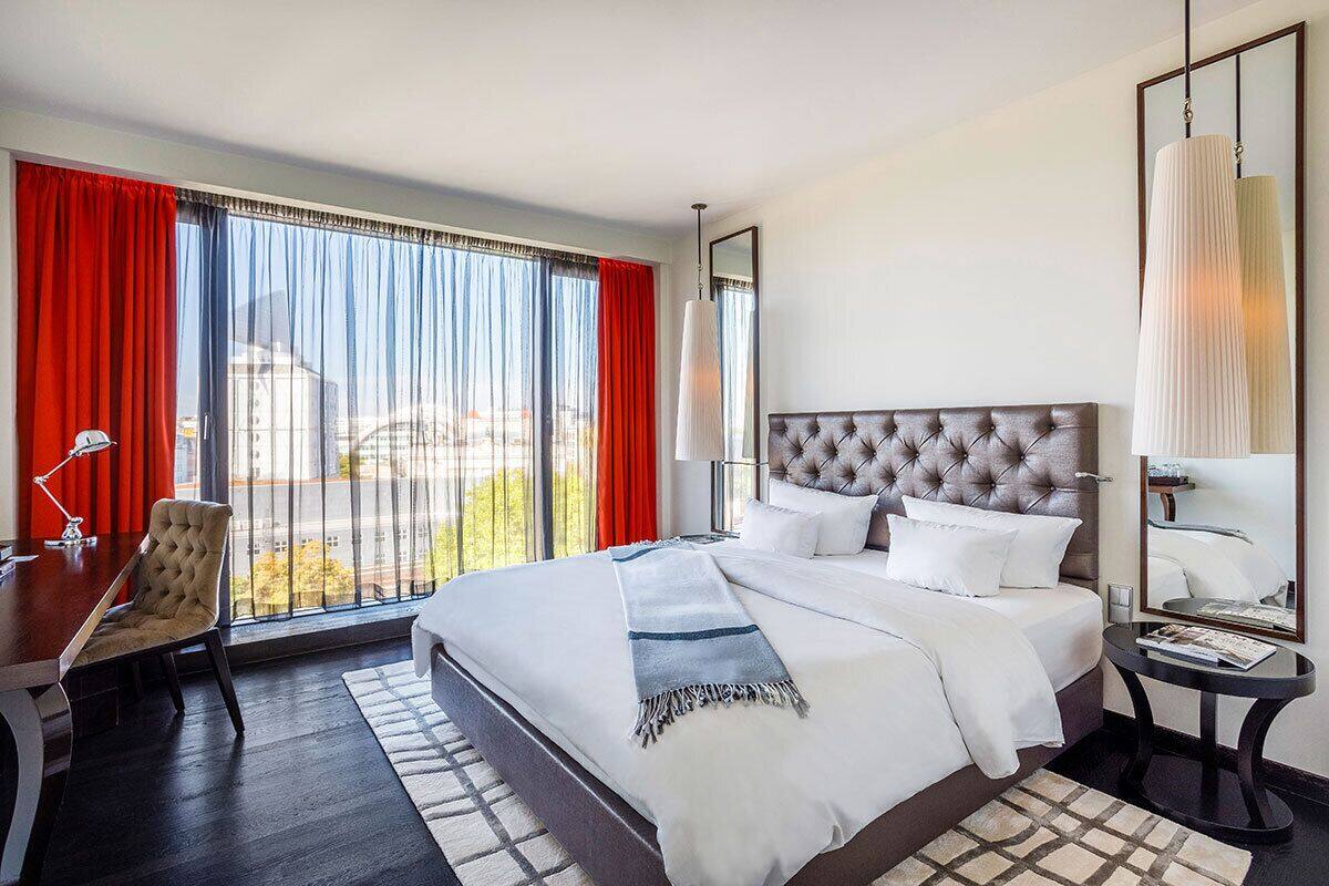 A hotel room with a large bed, a desk, and a chair. There is a window with curtains, a lamp, and a mirror on the wall.