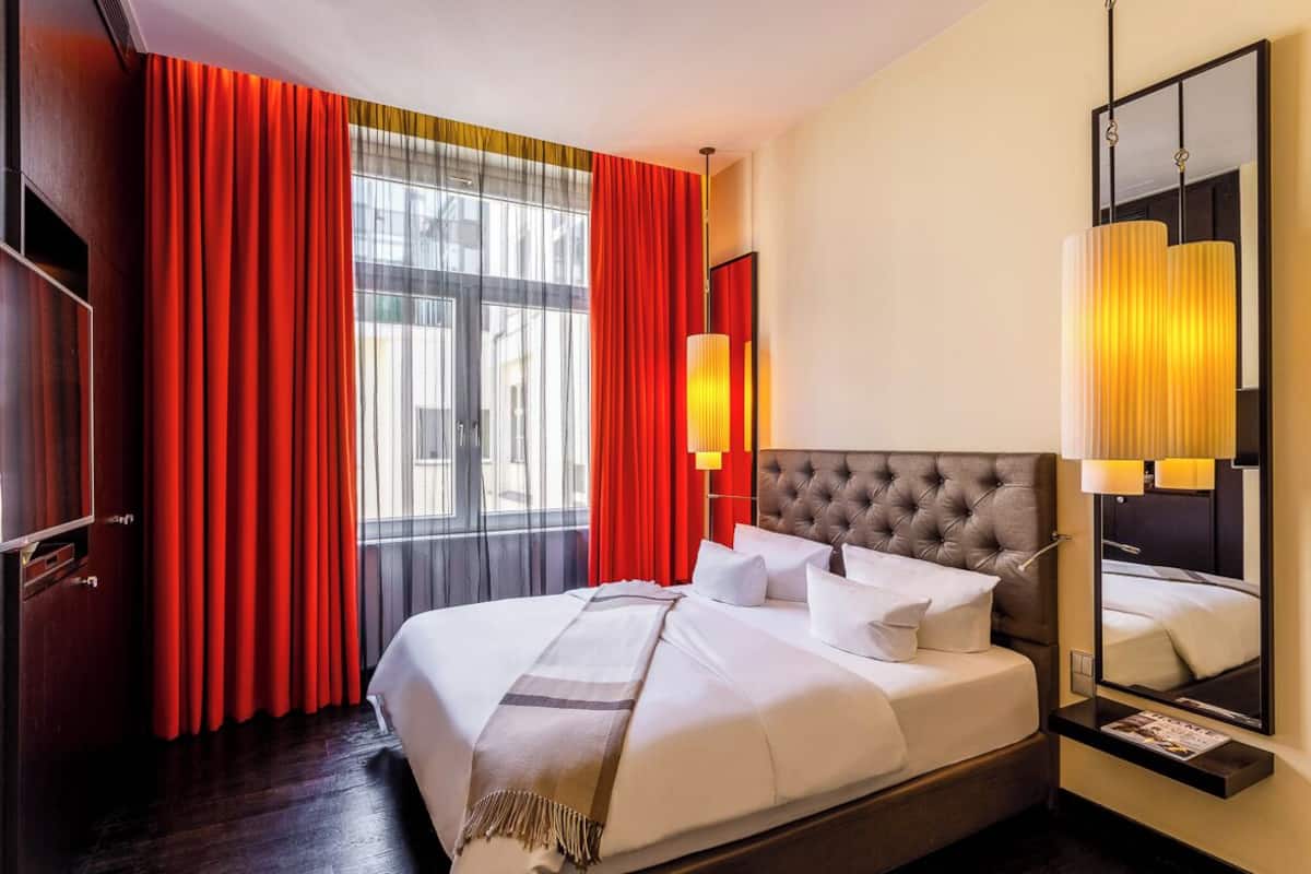 A hotel room with a large bed, red curtains, a large window, a wall-mounted mirror, and a bedside lamp.