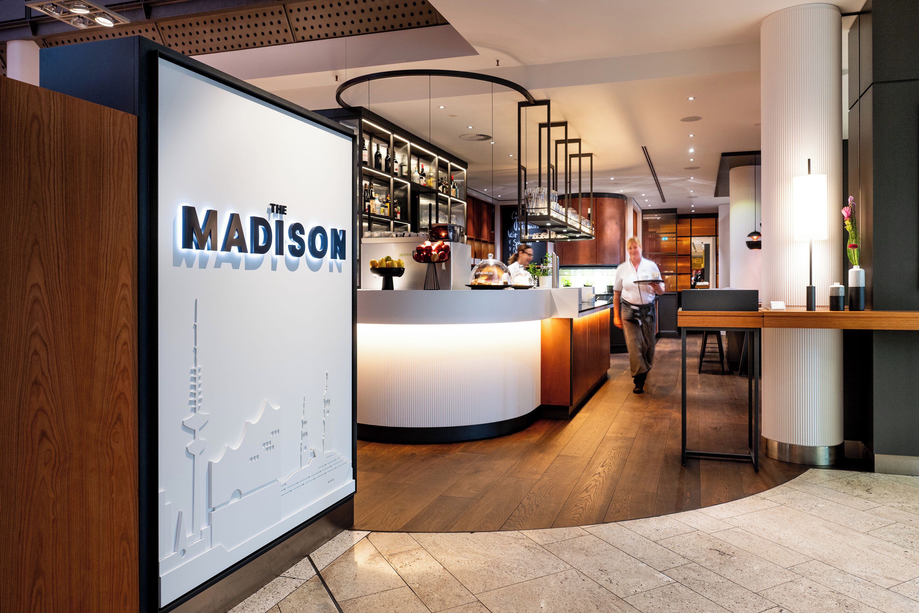 A modern hotel lobby with a reception desk, a bar area, and a large wall display.