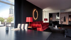 Una sala de estar moderna con un sofá rojo, una mesa de centro redonda con fruta y vistas a una torre del reloj a través de amplios ventanales.