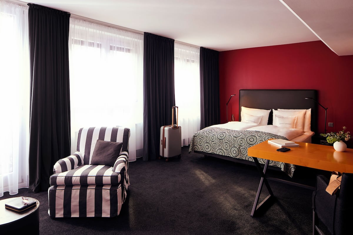 Een hotelkamer met een gestreept fauteuil, een bed met behang, een bureau met een lamp en een koffer.
