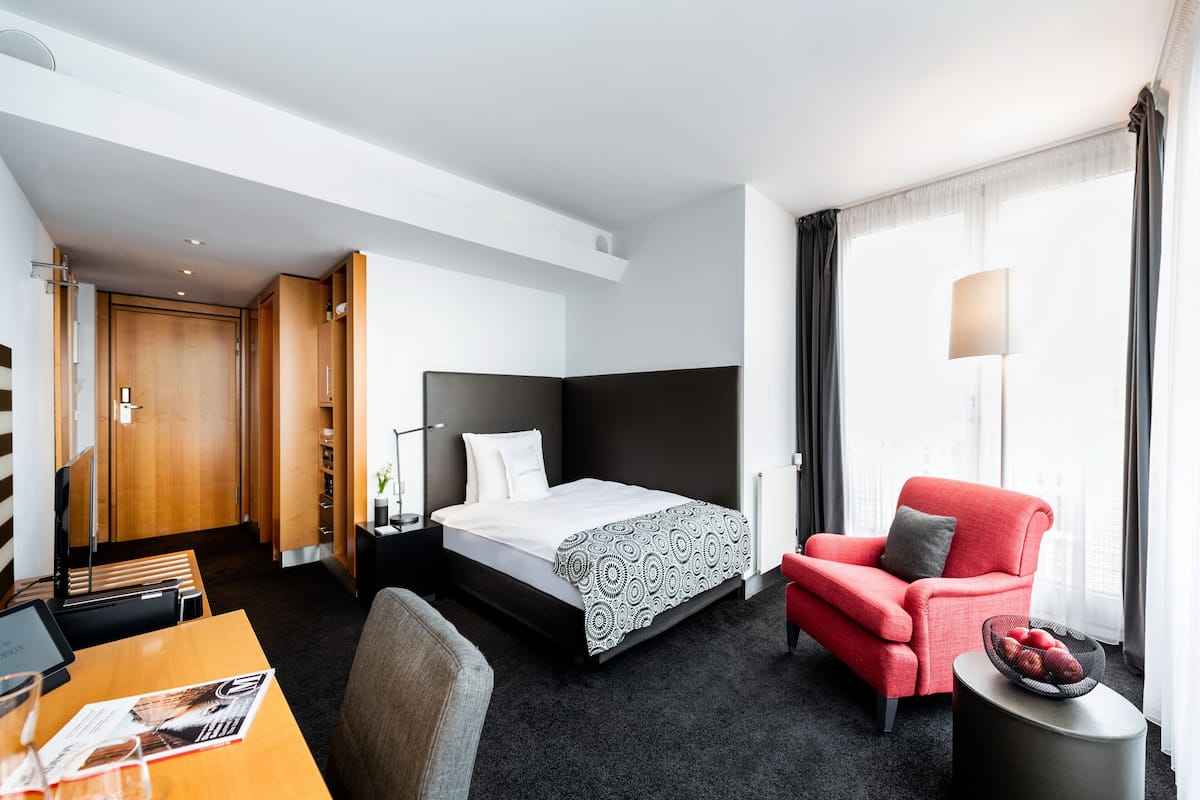 Een hotelkamer met een bed, een rode fauteuil, een tafeltje met een tijdschrift en een lamp.