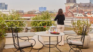 Una terraza en la azotea con una mesa, sillas y una persona de pie. Se observa una torre de reloj al fondo.