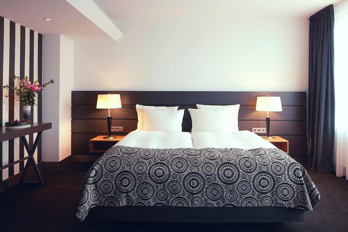 Een hotelkamer met een groot bed, twee nachtkastjes met lampen, een klein tafeltje met een bloemenvaas, en een donker hoofdeinde.