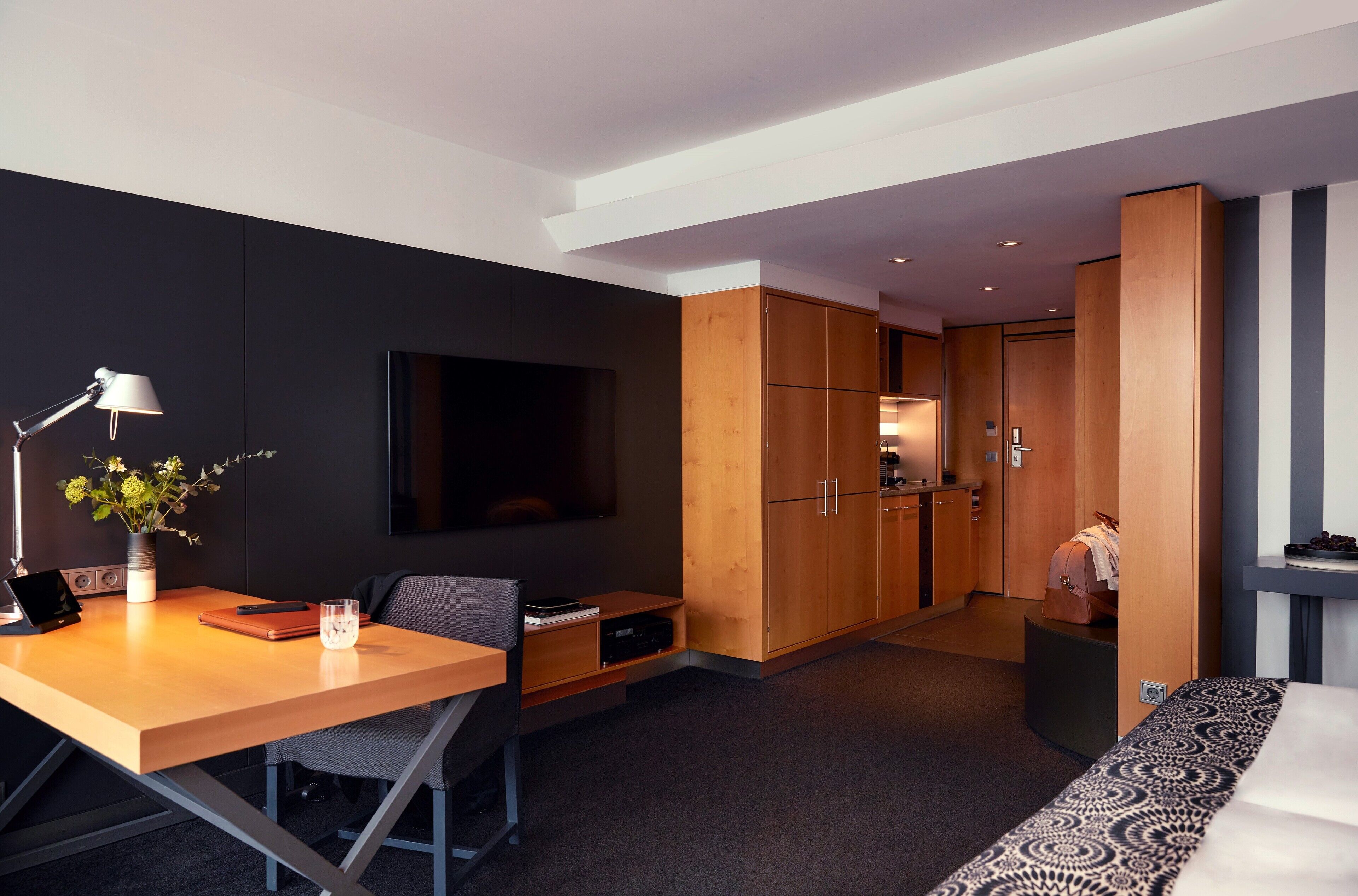 A modern hotel room with a wooden desk, a flat-screen TV, a small kitchenette, and a bed with patterned bedding.