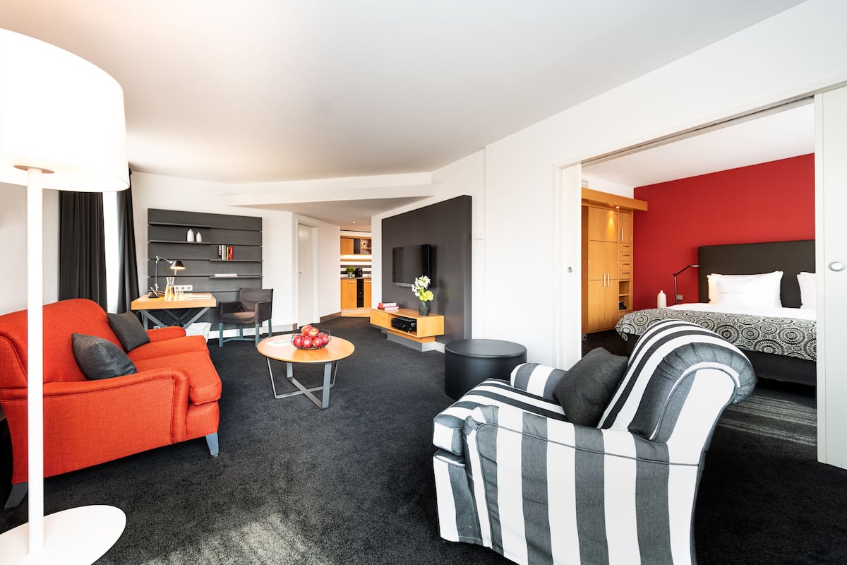 Een moderne hotelkamer met een rode accentmuur, een zwart-wit gestreepte bank, een rode bank, een zithoek met een tafel en stoelen, en een keuken op de achtergrond.