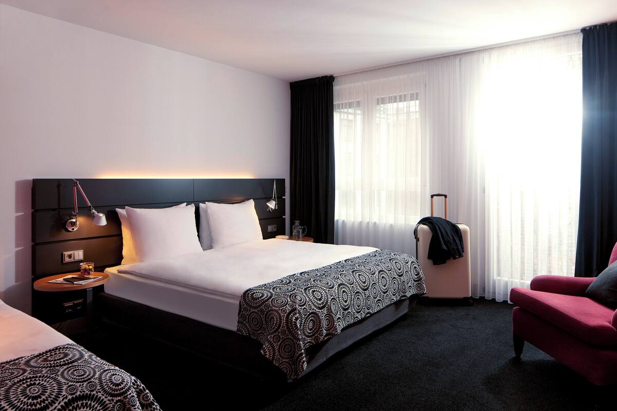 Een moderne hotelkamer met een groot bed, een nachtkastje met lamp, een stoel en uitzicht op de stad door dunne gordijnen.