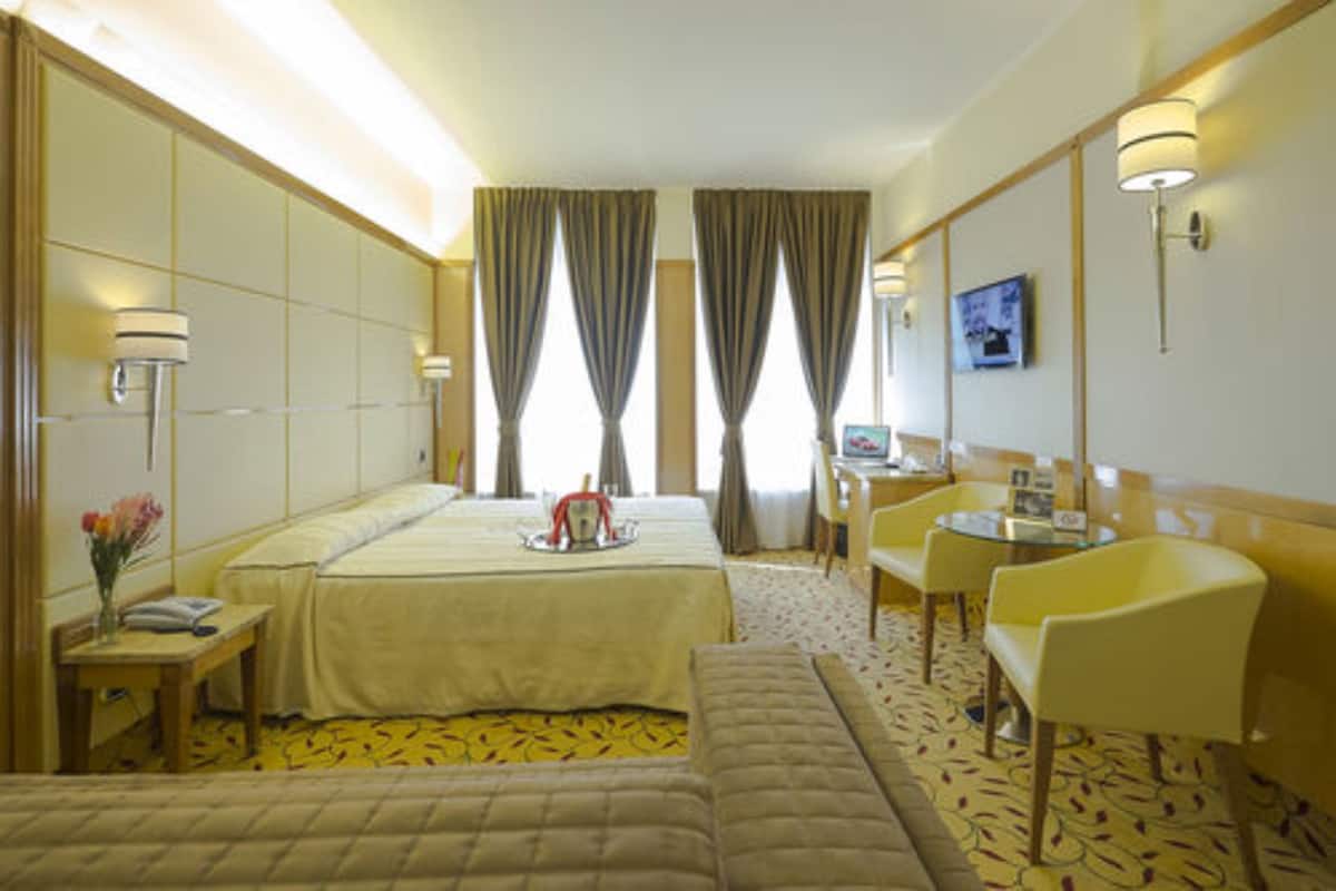 Une chambre d’hôtel comprenant un lit, une table de chevet, une lampe, une chaise et une petite table sur laquelle est posé un vase de fleurs.