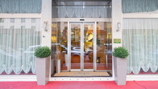 L’entrée d’un hôtel, avec une porte en verre, un tapis rouge et deux plantes en pot.