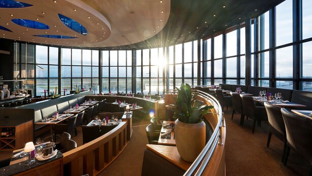 A restaurant with a view of the city skyline and the ocean. It features a bar area, multiple dining tables, and a large potted plant. - ATLANTIC Hotel Airport (Bremen)