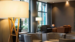 A modern lounge area with a large floor lamp, comfortable seating, and large windows offering a view of the outdoors. - ATLANTIC Hotel Airport (Bremen)