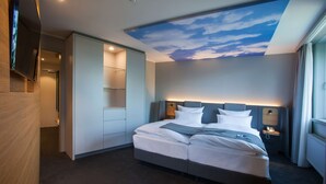 A modern hotel room with a large bed, a built-in TV, a ceiling with a sky mural, and a nightstand with a phone. - ATLANTIC Hotel Airport (Bremen)