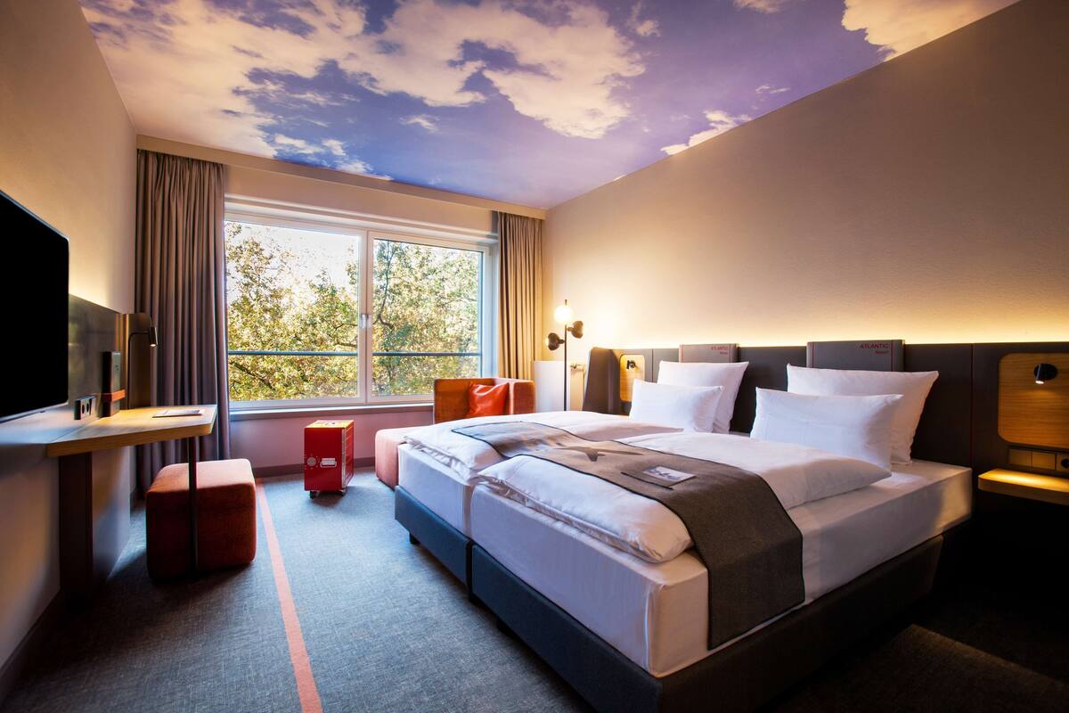 Una habitación de hotel con cama, escritorio, televisor y ventana con cortinas.