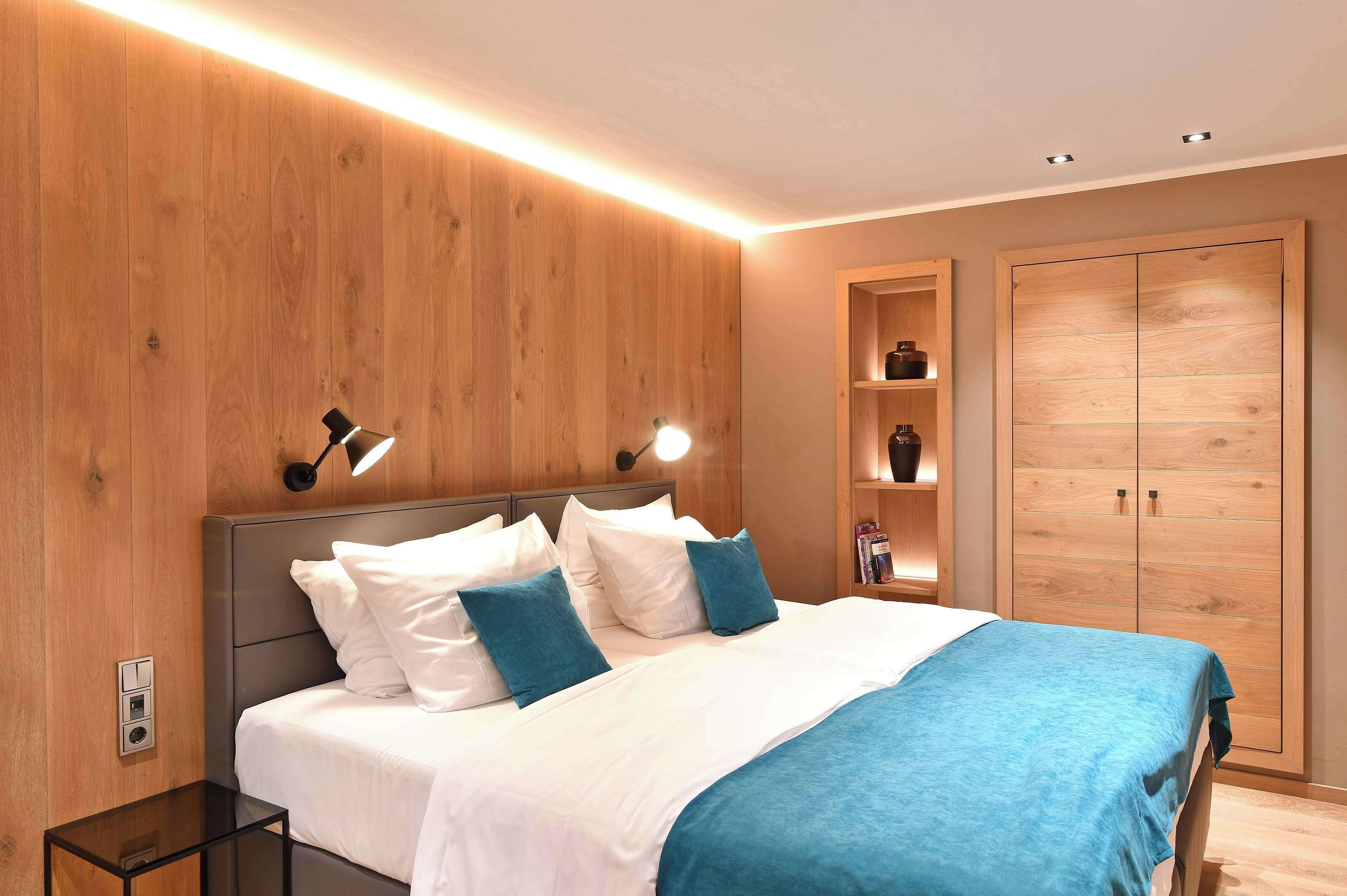 A modern bedroom with a wooden headboard, a bed with white and blue bedding, bedside lamps, a nightstand, and a wardrobe.