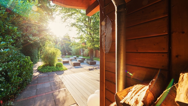 Une terrasse en bois avec un foyer, entourée de verdure et d’un bâtiment en bois.