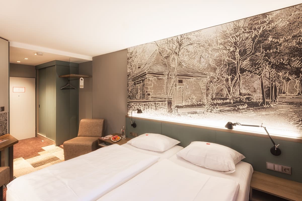 A hotel room with a large bed, a chair, a desk, and a large window with a view of a building and trees.