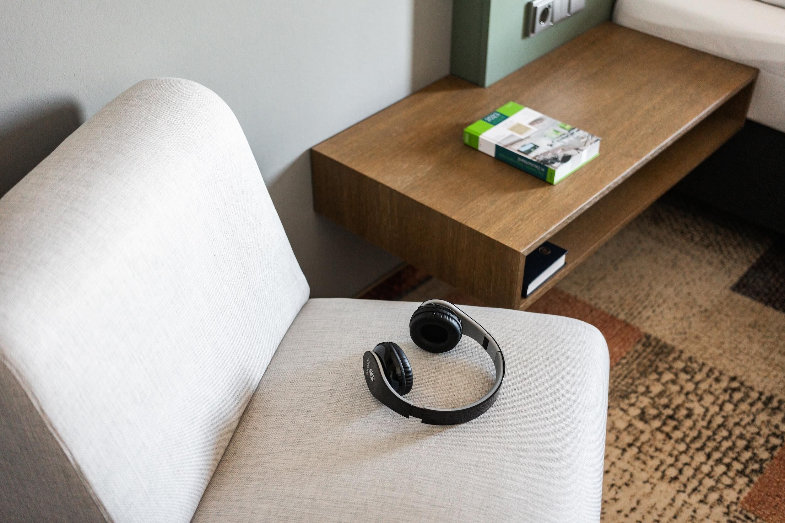 A white upholstered chair with a pair of black headphones on it, a wooden side table with a magazine, and a patterned rug on the floor.