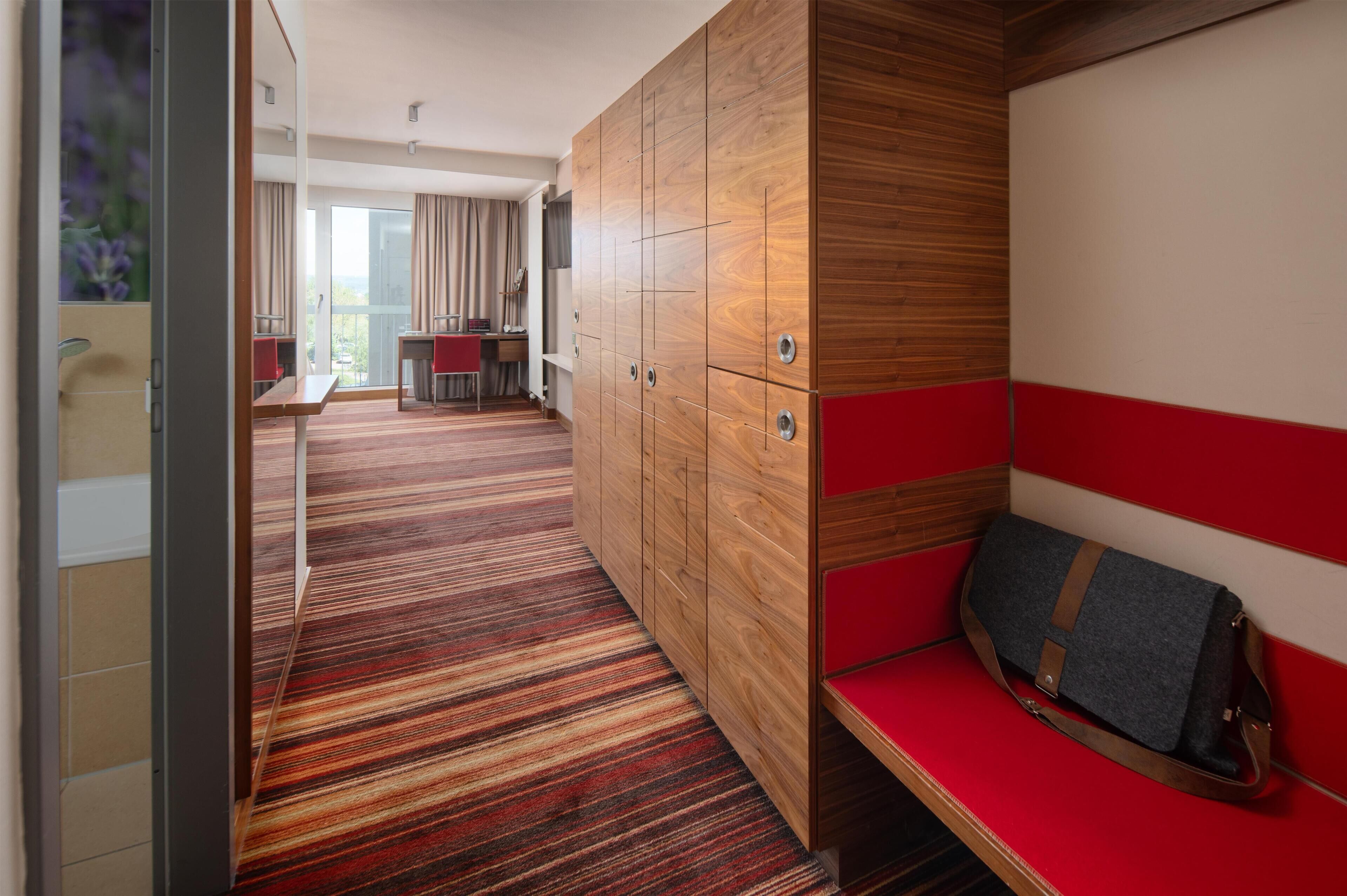 A hotel room with a wooden wardrobe, a red bench, and a striped carpet.
