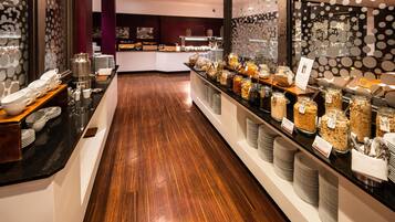 Un área de bufé con diversos alimentos, una barra con platos y cubiertos, y piso de madera.