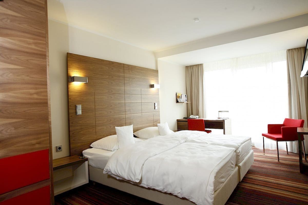 A hotel room with a large bed, a wooden headboard, a desk with a chair, and a window with curtains.