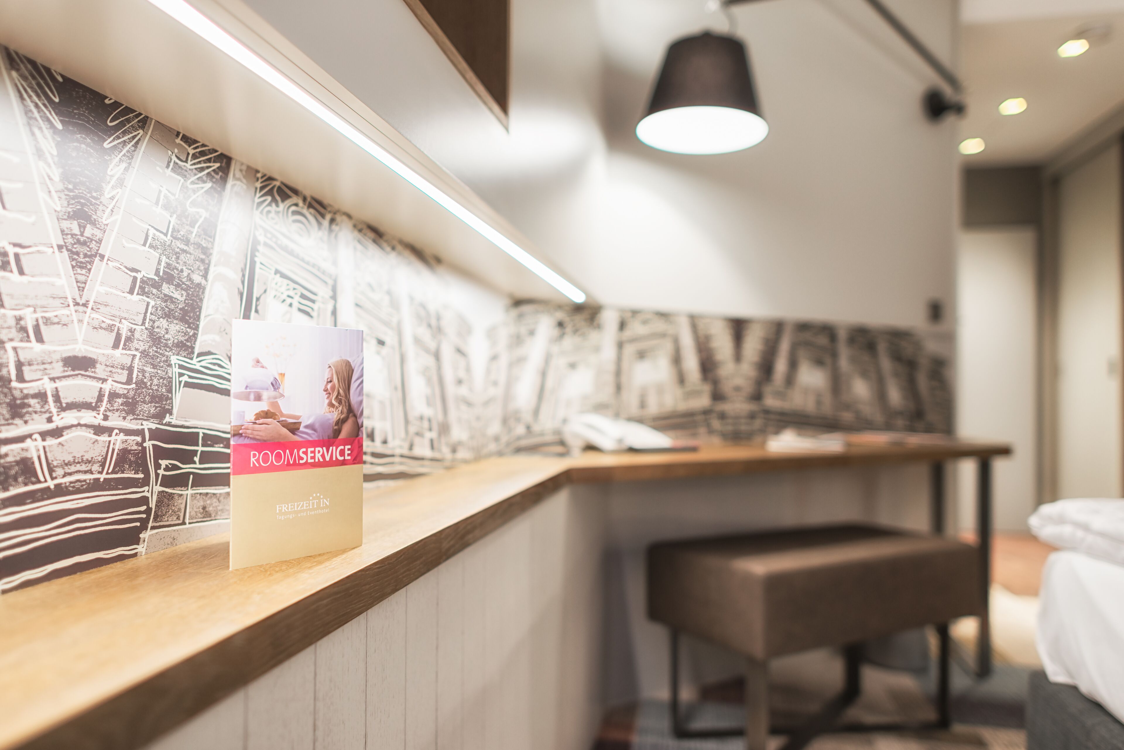 A hotel reception desk with a framed picture of a cityscape, a wooden counter, a bench, and a hanging light fixture.